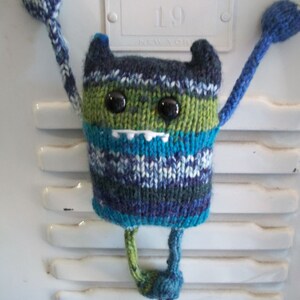 May include: A blue, green, and white knitted monster toy with googly eyes and a toothy grin. The toy has long arms and legs and is hanging from a white surface.