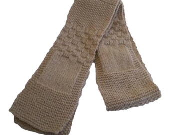 Può includere: Una lunga sciarpa lavorata a maglia di colore marrone chiaro che presenta una combinazione di texture. Il design include sezioni a legaccio, quadrati a maglia rasata liscia e un motivo a cestino in rilievo lungo tutto il tessuto.