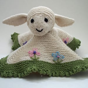 May include: A knitted white lamb with green and white blanket. The lamb has embroidered flowers on the blanket.