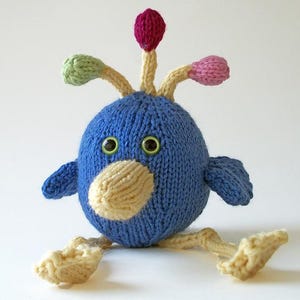 May include: A round, blue knitted creature toy with a cream-colored beak and two green eyes. It features three stalks on top with small, rounded tips in magenta, light green, and pink, along with two cream-colored legs and feet.