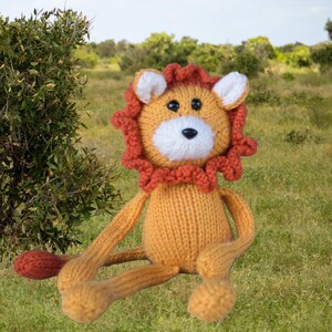 May include: A knitted lion stuffed animal featuring a golden yellow body, a white muzzle, and a dark orange mane. The toy has black plastic eyes, a black stitched nose, and a long tail with a dark orange tuft at the end, crafted from yarn.