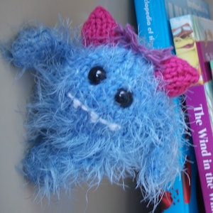 May include: A blue fuzzy knitted monster with pink ears and a white toothy grin. The monster is sitting on a shelf with books behind it.