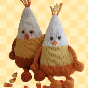 Puede incluir: Dos peluches de maíz dulce de punto con rayas naranjas, amarillas y blancas. Los peluches tienen ojos saltones y están sentados sobre un fondo a cuadros amarillo y blanco con maíz dulce esparcido.