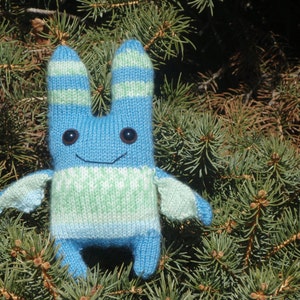 May include: A blue and green knitted plush toy shaped like a rabbit with a smiling face. The toy has a light blue body with green stripes and a white and green striped sweater.