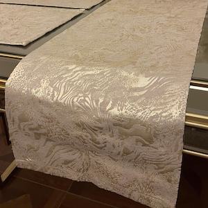 Peut inclure: Chemin de table beige avec un motif texturé et tourbillonnant. Le chemin de table est fait d'une matière douce, possiblement tissée. Le design comprend des nuances de crème et d'or, créant un aspect luxueux. Des sets de table assortis sont visibles.