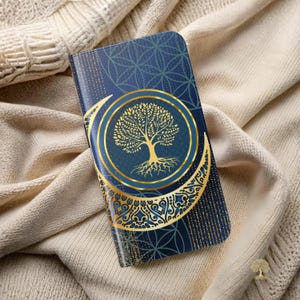 May include: A dark blue phone case with a gold tree of life design. The case features a gold crescent moon and circular accents. The background has a textured pattern with gold flecks, and the case is resting on a cream-colored knitted fabric.