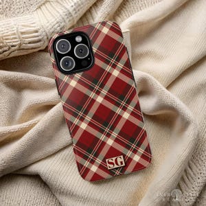 May include: A smartphone case featuring a diagonal red, cream, and dark green plaid pattern. The case has a black camera module cutout and a rectangular gold-toned emblem at the bottom centre displaying the capital letters SG in a serif font.