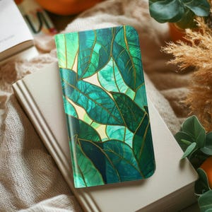 May include: A green and gold leaf pattern adorns a phone case. The case is resting on a beige book, with a soft, textured background. The design features various shades of green with gold accents, creating a vibrant and natural aesthetic.