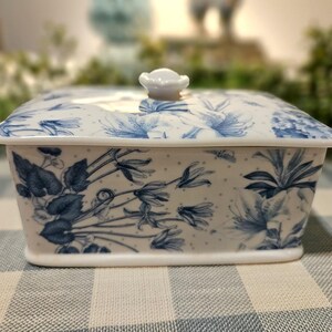May include: A rectangular porcelain butter dish with a white base and a blue floral pattern featuring delicate flowers and leaves. The lid includes a small, rounded knob handle on top, and the entire piece is decorated with a subtle blue dot pattern.