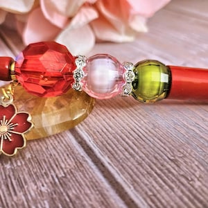 May include: A red pen with decorative beads and a cherry blossom charm. The pen features a faceted red bead, a pink bead, and a green bead, all accented with silver-colored spacers. A gold-colored cherry blossom charm hangs from the pen.