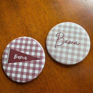 May include: Two round pinback buttons featuring a mauve and white gingham pattern. One button displays the word Bama in script, while the other features a maroon pennant graphic with the word Bama written in white script across the center.