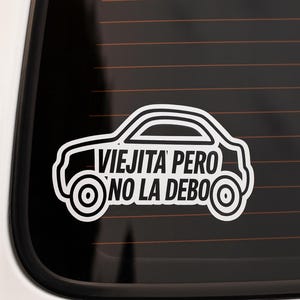 May include: White car sticker with the text "Viejita Pero No La Debo" in a black car window. The sticker features a car outline with wheels and a roof. The text is in a bold, sans-serif font.