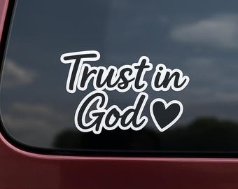 May include: White decal with the words "Trust in God" in a cursive font, with a heart symbol at the end. The decal is displayed on a dark background, possibly a car window.