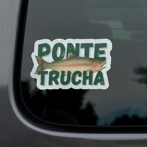 May include: A sticker showing a trout fish illustration with the words "PONTE TRUCHA" in green, outlined in white. The sticker is attached to a car window, with a blurred outdoor background visible.