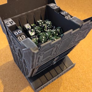 May include: A dark gray 3D-printed dice tower with a castle-like structure, featuring integrated mechanical counters displaying the numbers 00 and 99. The tower is filled with numerous small, dark green and black speckled dice with white pips.