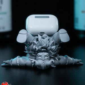 May include: A grey, stylised character figurine in a handstand position, supporting a white wireless earbud charging case on its feet. The character has spiky hair, a mask-like face, and muscular arms extended outwards. The base features the text PATREON h3LLcreator.