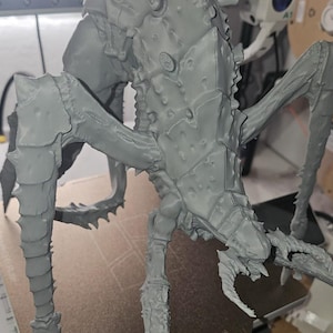 May include: A grey, unpainted 3D printed figurine of a multi-legged creature with a segmented, armoured exoskeleton. The model features sharp, pointed protrusions along its limbs and a detailed, textured carapace with a complex, insectoid design.