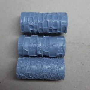 May include: Three cylindrical grey rollers featuring distinct embossed textures on their surfaces. The top roller has a grid pattern, the middle roller displays a brick-like design, and the bottom roller shows a cracked, irregular stone-like texture.