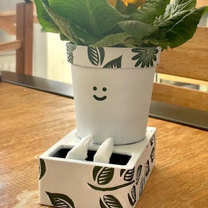 Puede incluir: Una maceta de cerámica blanca con un patrón de hojas verdes pintado a mano y una cara sonriente negra sencilla. La maceta cilíndrica se asienta sobre una base rectangular, con dos pequeños pies de cerámica blanca en la parte frontal.