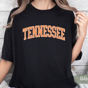 May include: Black t-shirt with the word "TENNESSEE" in orange block letters. The letters have a checkered pattern. The shirt is short-sleeved and appears to be made of a soft material.