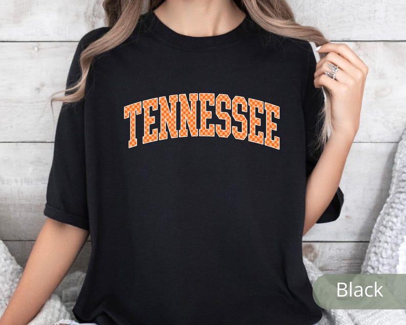 May include: Black t-shirt with the word "TENNESSEE" in orange block letters. The letters have a checkered pattern. The shirt is short-sleeved and appears to be made of a soft material.
