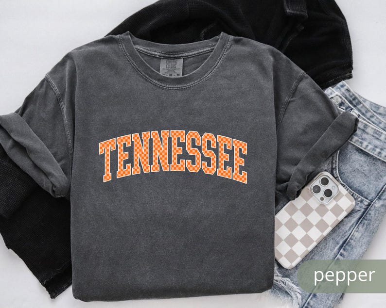 May include: A dark gray sweatshirt with the word "TENNESSEE" in orange and white checkered letters. The sweatshirt is folded with a pair of jeans and a phone case with a checkered pattern. The word "pepper" is in the bottom right corner.