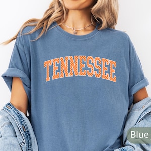 May include: A blue t-shirt with the word "Tennessee" in orange block letters. The letters have a checkered pattern. The shirt has rolled-up sleeves and a relaxed fit.