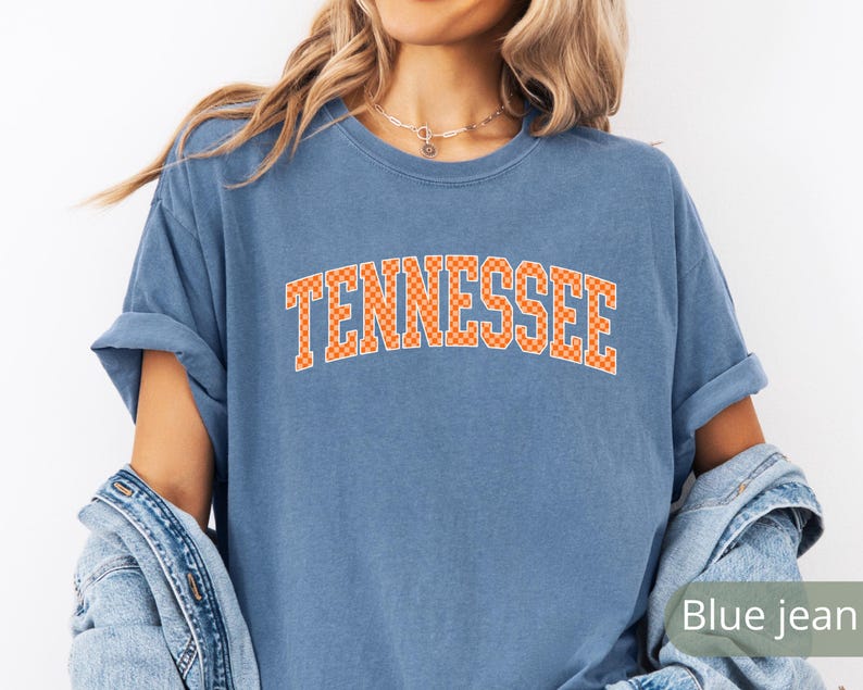 May include: A blue t-shirt with the word "Tennessee" in orange block letters. The letters have a checkered pattern. The shirt has rolled-up sleeves and a relaxed fit.