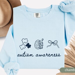 Puede incluir: Sudadera azul claro con las palabras "concienciación sobre el autismo". El diseño incluye piezas de rompecabezas, un cerebro y un lazo. La sudadera es de un material suave y de ajuste cómodo.