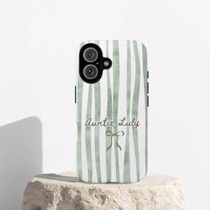 May include: A white phone case with a vertical green stripe pattern. The text "Auntie Luly" is written in a script font, with a small bow graphic below it. The phone case is on a stone surface.