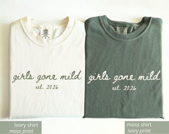 May include: Two folded t-shirts, one ivory and one moss green, each with the phrase "girls gone mild" and the year "est. 2026" printed on the front. The ivory shirt has a moss green print, and the moss shirt has an ivory print.