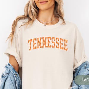 May include: Ivory-colored t-shirt with the word "TENNESSEE" in orange checkered letters. The shirt has rolled-up sleeves and is paired with a denim jacket. The overall style is casual and relaxed.