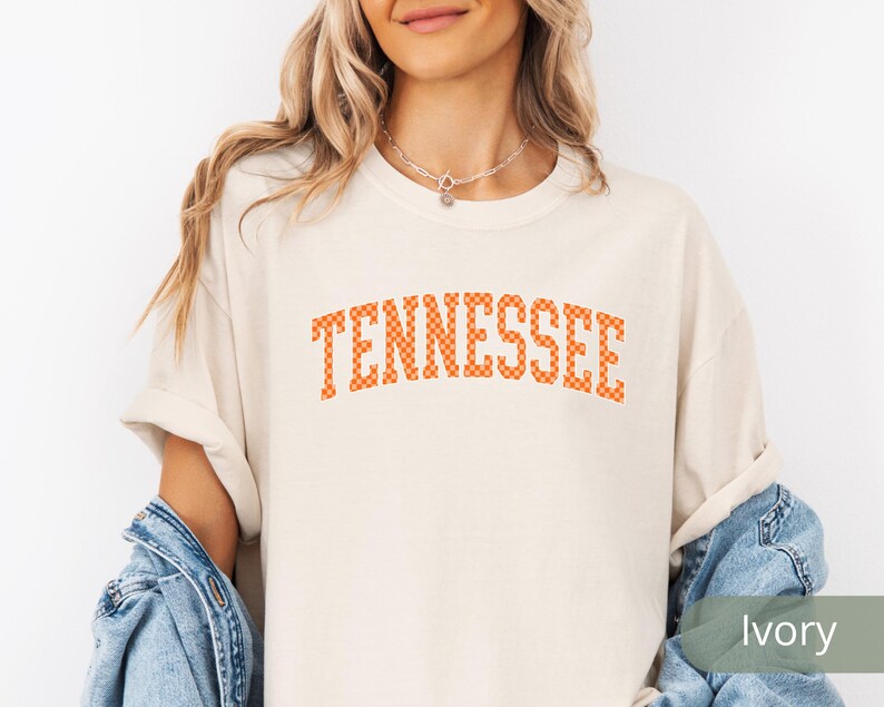 May include: Ivory-colored t-shirt with the word "TENNESSEE" in orange checkered letters. The shirt has rolled-up sleeves and is paired with a denim jacket. The overall style is casual and relaxed.