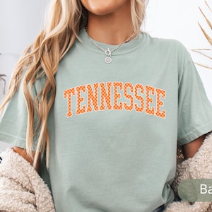 May include: A light green t-shirt with the word "TENNESSEE" in orange block letters. The letters have a checkered pattern. The shirt is a short-sleeved, crew neck style.
