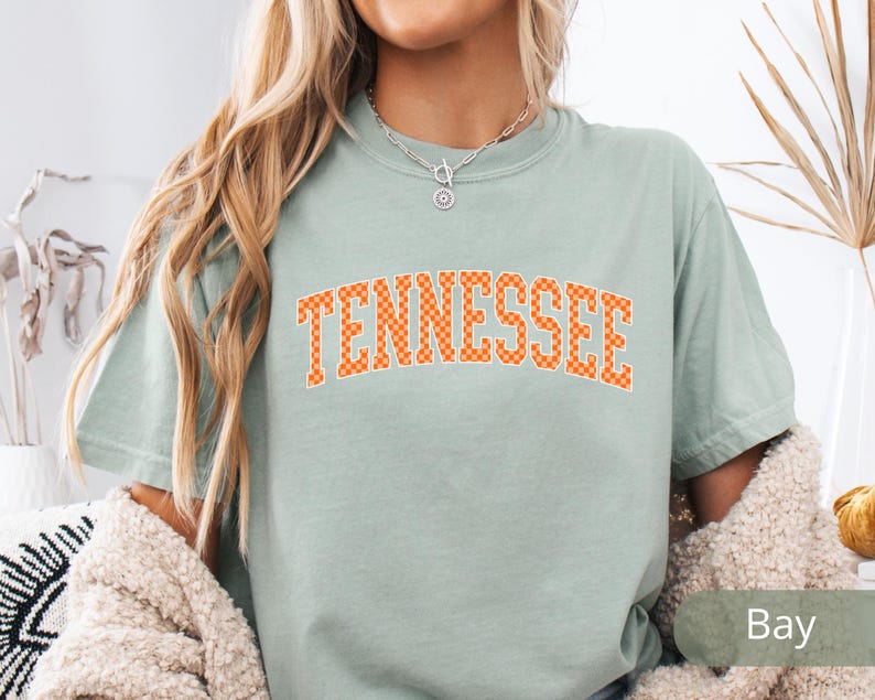 May include: A light green t-shirt with the word "TENNESSEE" in orange block letters. The letters have a checkered pattern. The shirt is a short-sleeved, crew neck style.