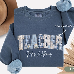 May include: A denim blue sweatshirt with the word "TEACHER" in a faux patchwork design. The patchwork includes floral and gingham patterns. The name "Mrs. Williams" is written below the word "TEACHER".