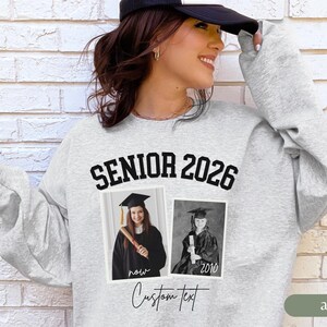 May include: Ash gray sweatshirt with the text "SENIOR 2026" in black block letters. Two framed photos of a graduate are below the text. The words "now" and "2010" are below the photos. The words "Custom Text" are written in cursive.