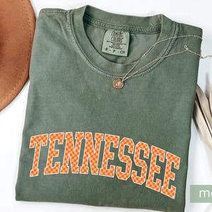 May include: A moss green t-shirt with the word "Tennessee" in orange checkered letters. A gold necklace with a pendant rests on the shirt. A brown hat and dried leaves are also in the image. The shirt has a Comfort Colors tag.