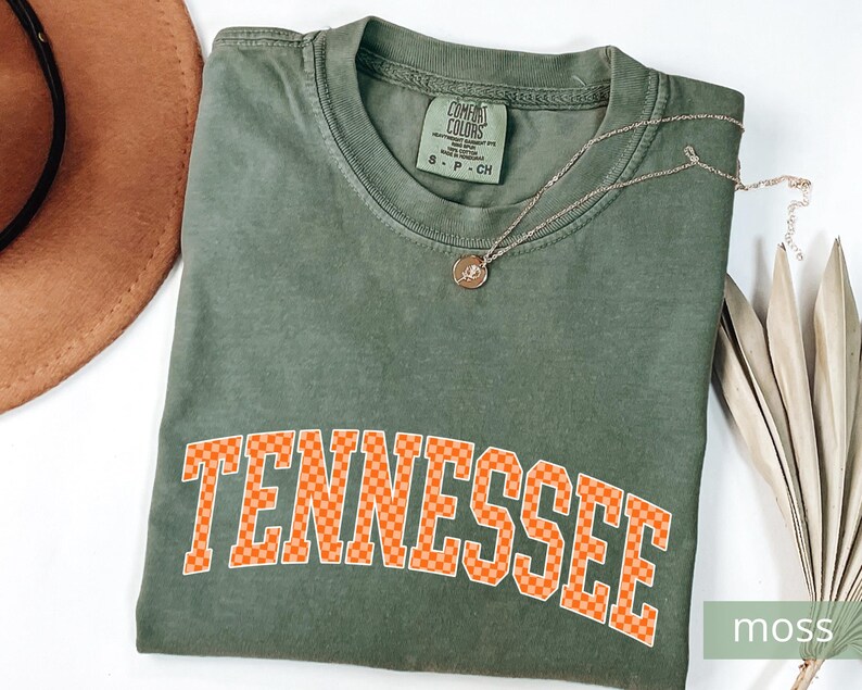 May include: A moss green t-shirt with the word "Tennessee" in orange checkered letters. A gold necklace with a pendant rests on the shirt. A brown hat and dried leaves are also in the image. The shirt has a Comfort Colors tag.