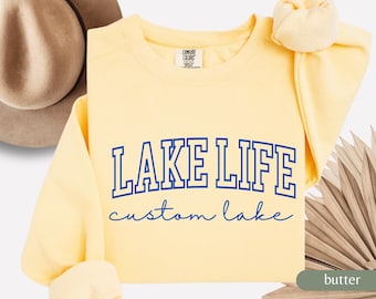 May include: A butter-yellow sweatshirt with the words "LAKE LIFE" in blue block letters and "custom lake" in a script font. The sweatshirt has a crew neck and long sleeves. A brown hat is visible in the background.