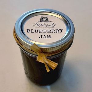 May include: A jar of blueberry jam with a silver lid and a paper label. The label reads "Propinquity WASHINGTON, MISSISSIPPI BLUEBERRY JAM". A raffia bow is tied around the jar. The jam is dark purple.