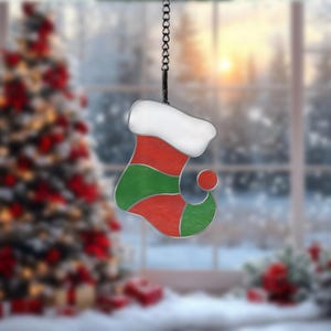 Puede incluir: Adorno navideño de calcetín de vidriera. El adorno presenta un diseño rojo y verde con un puño blanco. Cuelga de una cadena negra. El fondo es una escena invernal borrosa con un árbol de Navidad.