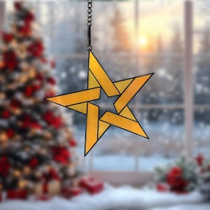 May include: A hanging star-shaped ornament with a geometric design. The star is made of yellow glass with black outlines and is suspended by a chain. The background shows a Christmas tree and a snowy window scene.