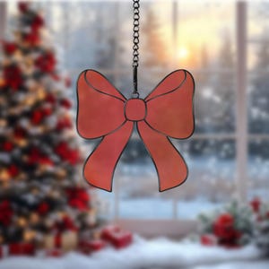 May include: A red stained glass bow ornament hanging from a chain. The ornament is in front of a blurred background of a Christmas tree and a window.