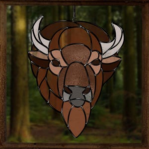 May include: A stained glass window hanging features a brown bison head with white horns. The bison is set against a blurred background of a forest.