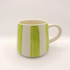 Könnte beinhalten: Eine Keramik-Tasse mit cremefarbenem Innenraum und Henkel. Die Außenseite hat vertikale Streifen in leuchtendem Grün und Creme. Die Tasse ist zylindrisch mit rundem Boden und einem einfachen, gebogenen Henkel.