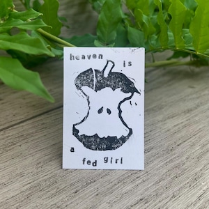 May include: A rectangular white card featuring a black ink block print of an apple core. The text "heaven is" is printed above the apple core, and the text "a fed girl" is printed below the apple core in a simple, sans-serif black font.