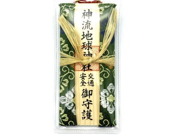 May include: A Japanese omamori charm in a clear plastic case. The charm is wrapped in green fabric with a white floral pattern and tied with natural-colored raffia. Japanese text is printed on the front.