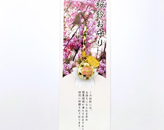 Op de afbeelding: Een Japanse charme met een goud- en roze bloemenontwerp, hangend aan een kleurrijk koord. De charme is bevestigd aan een witte kaart met Japanse tekst en een achtergrondafbeelding van roze kersenbloesems.
