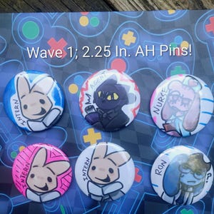 May include: Six circular pin-back buttons featuring cartoon character illustrations. The text 'Wave 1; 2.25 In. AH Pins!' is visible. The pins depict characters labeled 'Intern', 'Agent', 'Nurse', and 'Ron' with various colors and line art styles.